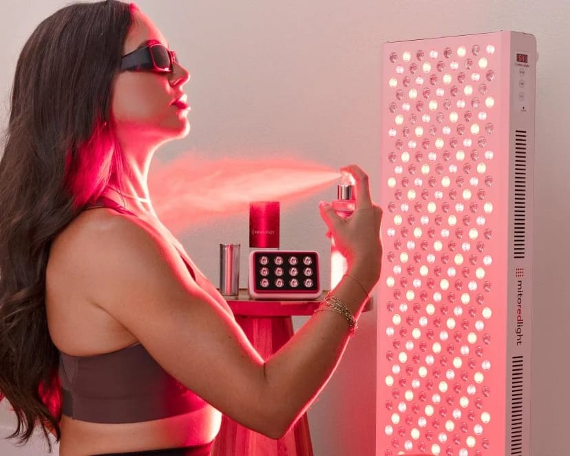 Woman spraying skincare onto her skin next to a Mito red light panel before a session