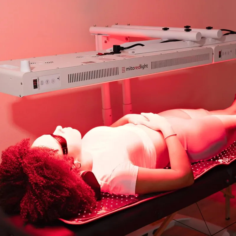 Woman lying on a treatment bed under a MitoPRO 1500+ red light panel mounted horizontally on its stand in a clinic