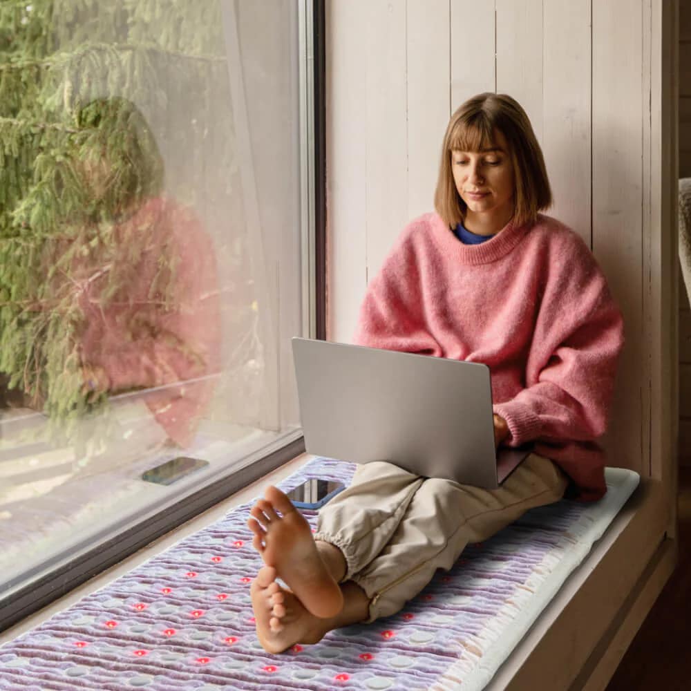 Woman working on laptop while seated on HealthyLine TAJ PEMF Mat for combined productivity and therapy