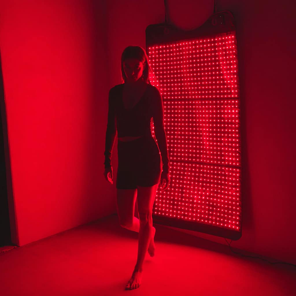 Woman using HigherDose Mat mounted on wall as standing LED therapy panel for vertical treatment