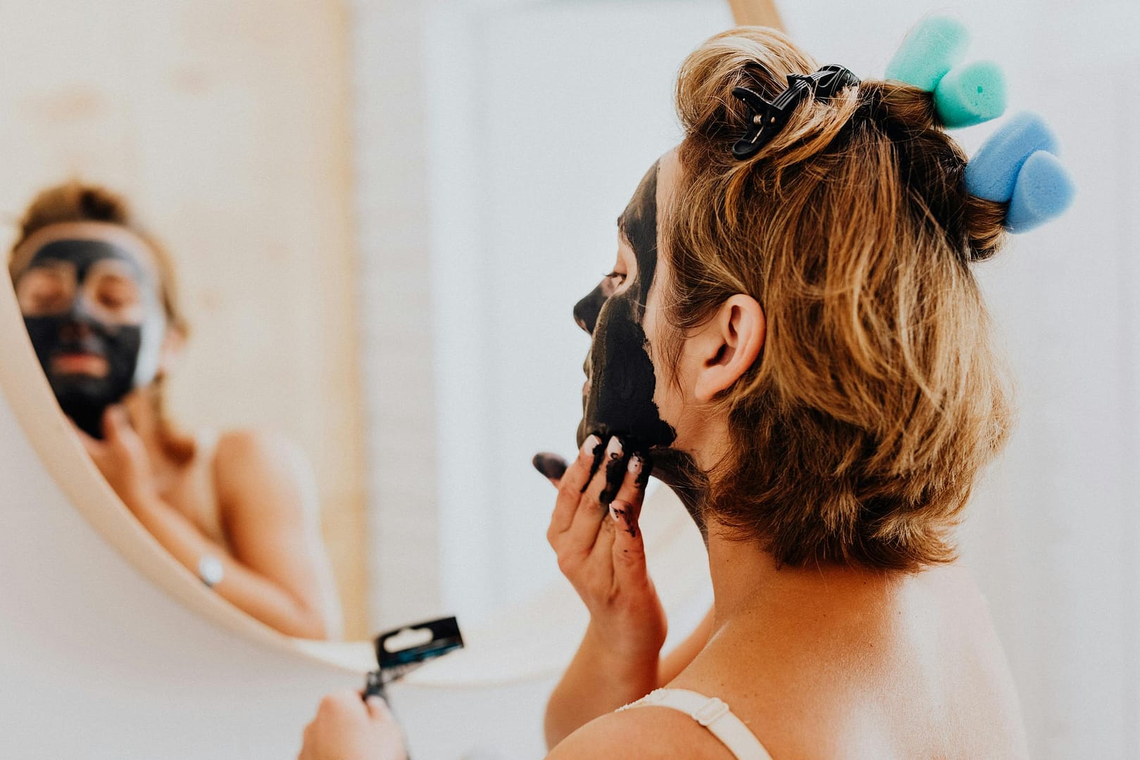 Red Light Therapy for Skin Care: A woman is applying a black facial mask in front of a mirror, focusing on skincare and beauty routines.