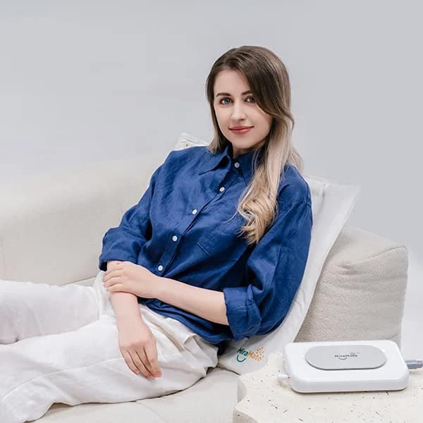 Woman using PEMF therapy pad with multiple coils for back pain relief on sofa