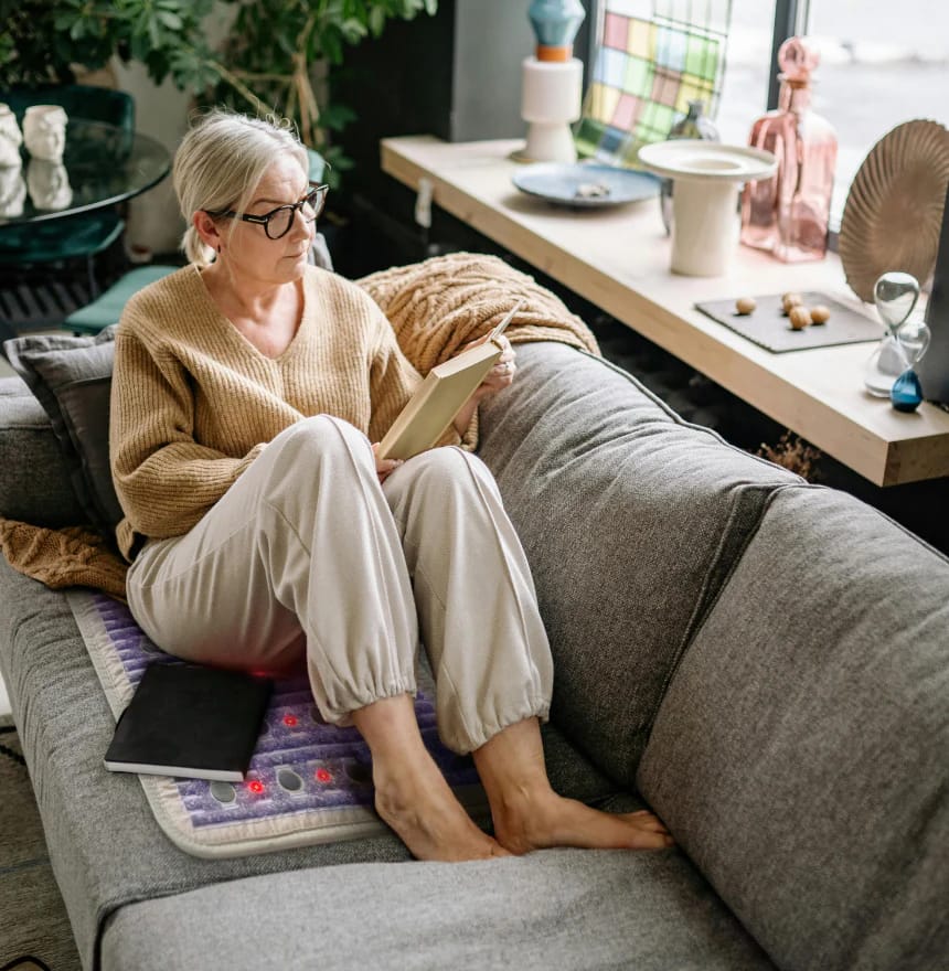 Woman relaxing on HealthyLine TAJ PEMF MiniMat on couch, enjoying PEMF therapy and reading